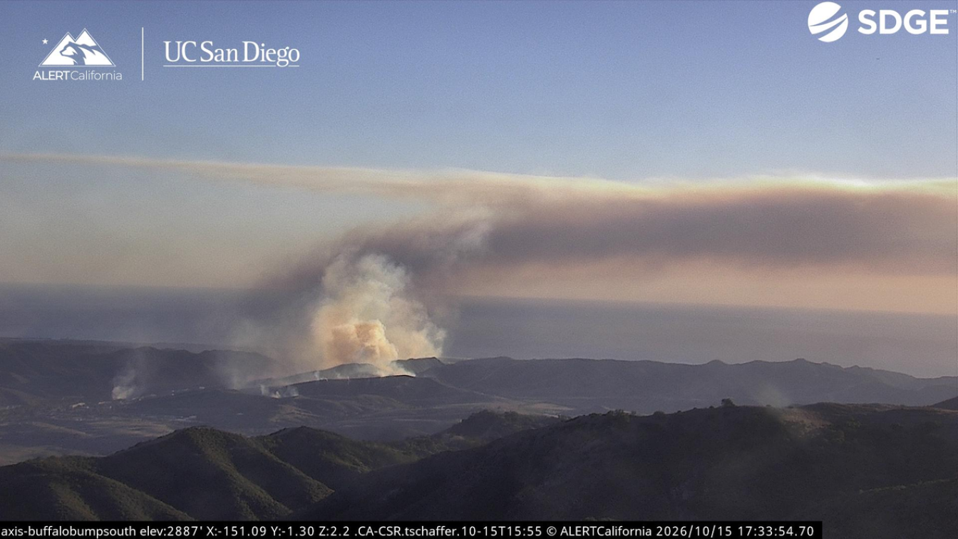 Smoke rises above a hilly landscape with the ocean in the background.