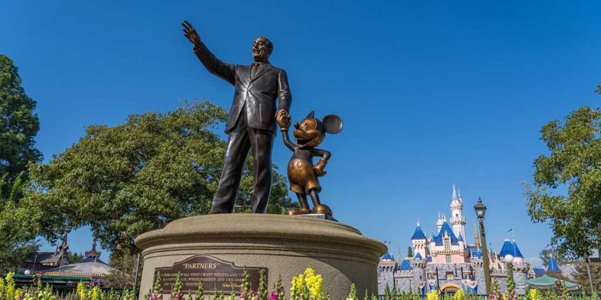 A bronze statue depicting Disney Legend holding hands with Mickey Mouse stands on a pedestal surrounded by flowers. The man is extending his right arm forward; in the background, there is a blue and white castle under a clear blue sky.